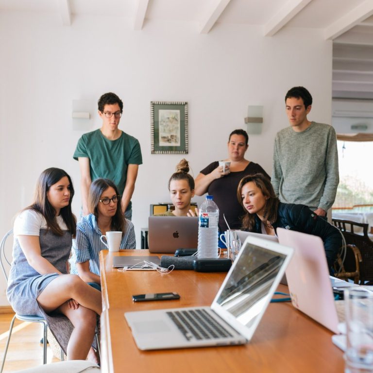 Un groupe de jeunes adultes sont regroupés autour d'un bureau et regardent ensemble l'ordinateur d'une personne.